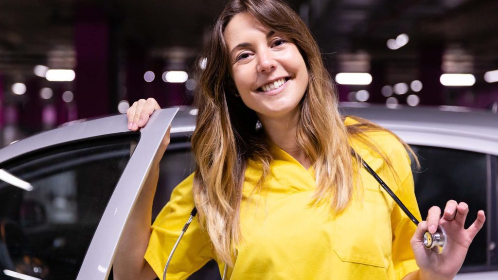 A brunette travel nurse stepping out of her silver car while holding a stethoscope and a car door, wearing a yellow scrub with a smile, representing mobility and readiness to provide medical care